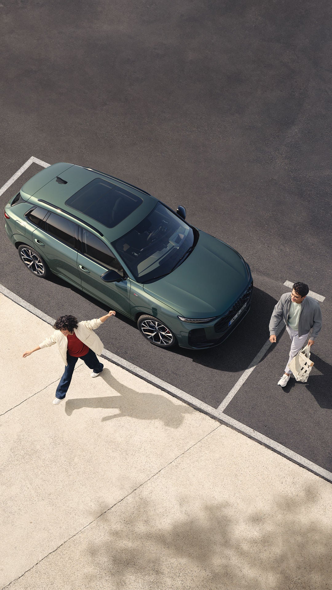 Une vue aérienne montre une Audi Q3 verte garée sur un parking. Deux personnes, un homme et un enfant, se tiennent debout près de l'avant du véhicule.
