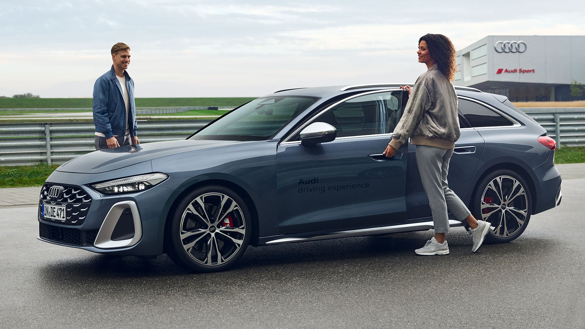 Ein grauer Audi Q3 SUV ist auf einer Rennstrecke geparkt, auf dessen Tür das Logo „Audi driving experience“ prangt. Zwei Personen sind zu sehen: ein Mann, der in der Nähe der Front steht, und eine Frau, die die Wagentür öffnet. Im Hintergrund sind das Gebäude von Audi Sport und ein Teil der Rennstrecke zu sehen.