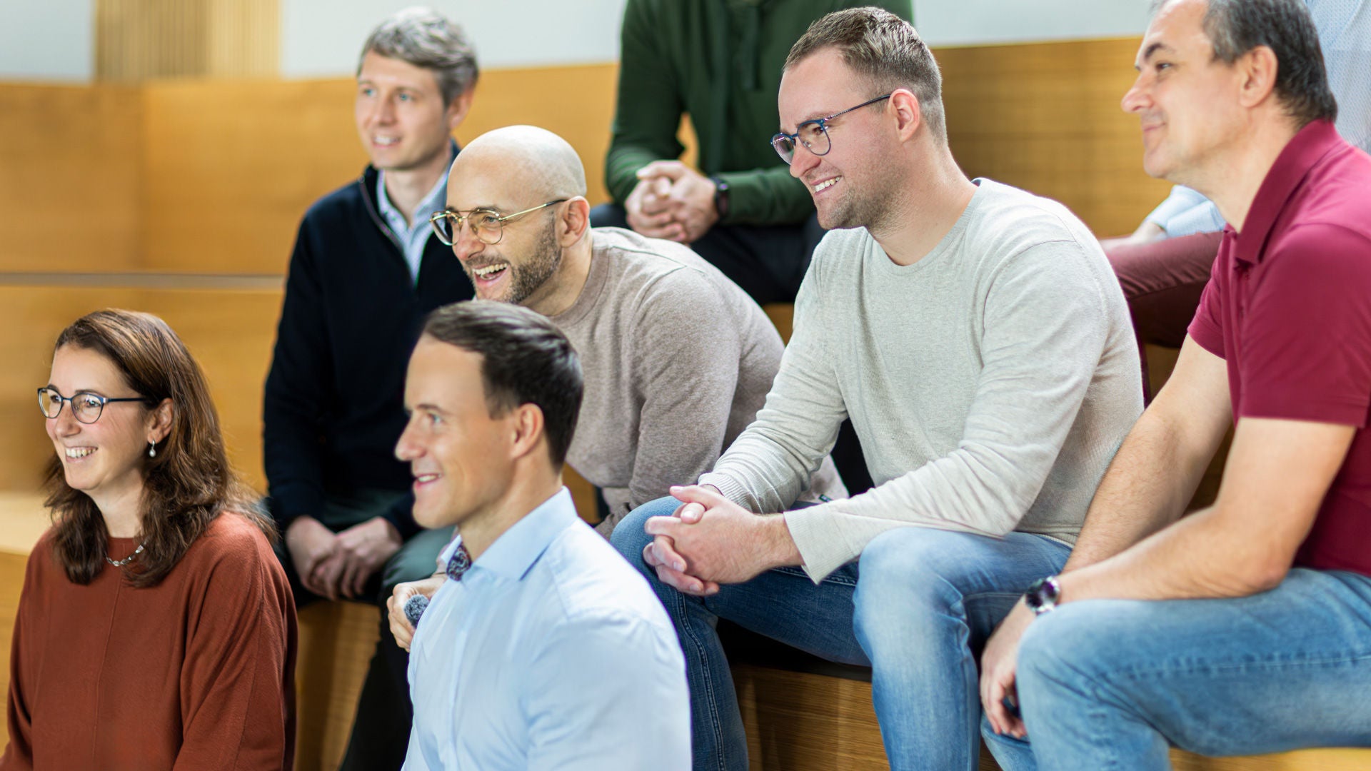 A group of Audi employees sitting on wooden steps in a bright room and looking ahead together.
