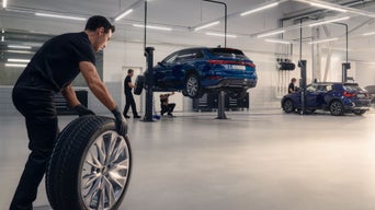 Mehrere Mechaniker arbeiten in heller Werkstatt an angehobenen Audi-Fahrzeugen, ein Mann rollt einen Reifen.