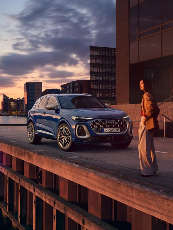 Ein blauer Audi SQ5 SUV parkt bei Sonnenuntergang an einem urbanen Kai, eine Frau steht daneben und blickt in die Ferne.
