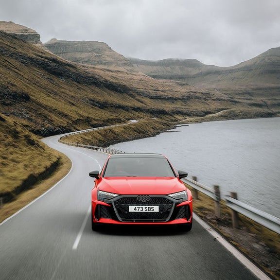A red RS 3 Sportback car drives along a winding lakeside road with rugged, grassy hills. The scene conveys excitement and adventure in a serene, natural setting.
