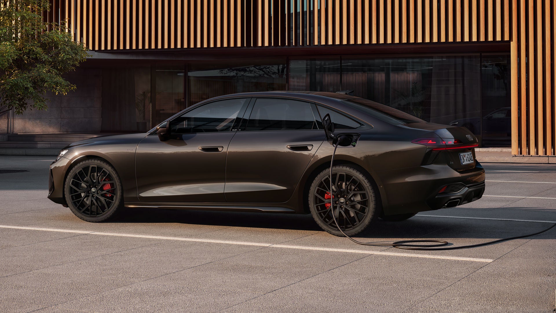 The A6 Saloon e-hybrid in Havana Black Metallic is parked sideways in front of a building facade and is connected to a charging station.