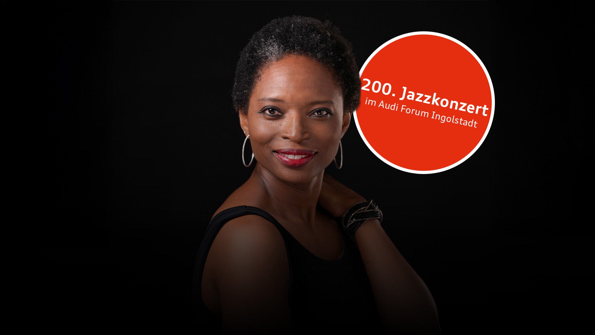 Porträt einer lächelnden Frau mit kurzem Haar, roten Lippen und Creolen; schwarzes Oberteil vor dunklem Hintergrund.