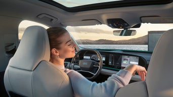 Frau sitzt entspannt mit geschlossenen Augen im Audi SUV Q6 e-tron, mit Blick auf Meer und Sonnenuntergang durch die Frontscheibe.