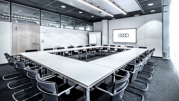Heller Konferenzraum mit U-förmigem Tisch, schwarzen Stühlen und Audi-Logo auf einem Bildschirm.