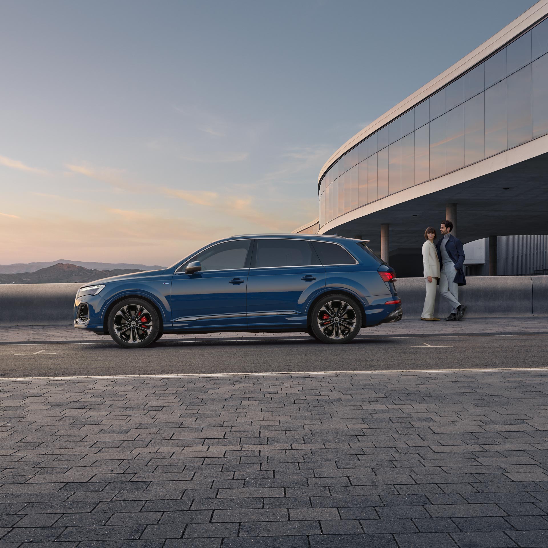 Ein blauer Audi Q7 SUV seitlich vor moderner Fassade, Paar im Businesslook steht lächelnd im Hintergrund.