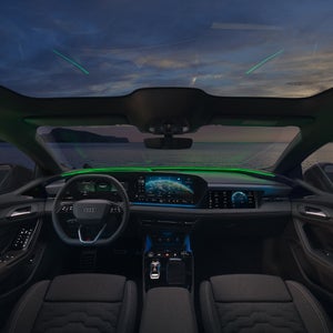 Audi Innenraum mit grünem Ambientelicht und digitalen Displays, im Hintergrund modernes Haus mit Treppe, Hecke und Sonnenuntergang.