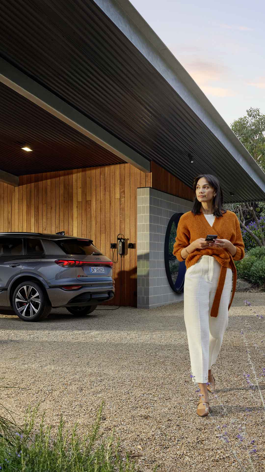 Dunkelhaarige Frau mit Smartphone läuft vor einem geparkten, ladenden Audi Q6 SUV e-tron unter Carport mit Holzfassade bei Tageslicht.