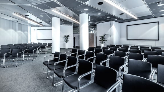 Raum mit Reihenbestuhlung, schwarzen Stühlen und zwei großen weißen Projektionsflächen.