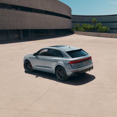 Ein grauer Audi RS Q8 SUV performance aus schräger Heckansicht auf heller Betonfläche vor moderner Architektur und Palmen bei Sonnenschein.