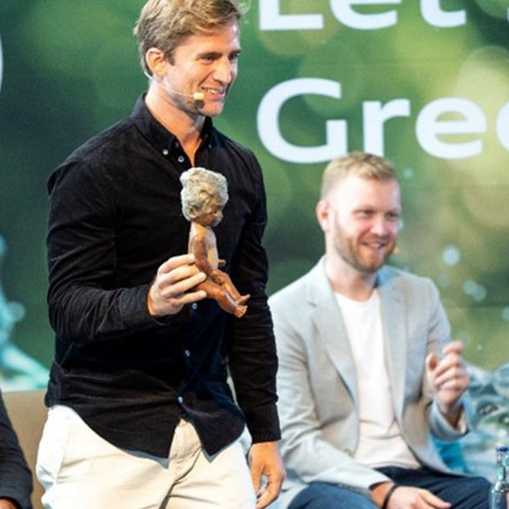 Man holding a doll on a stage. The Greenovation logo can be seen in the background.