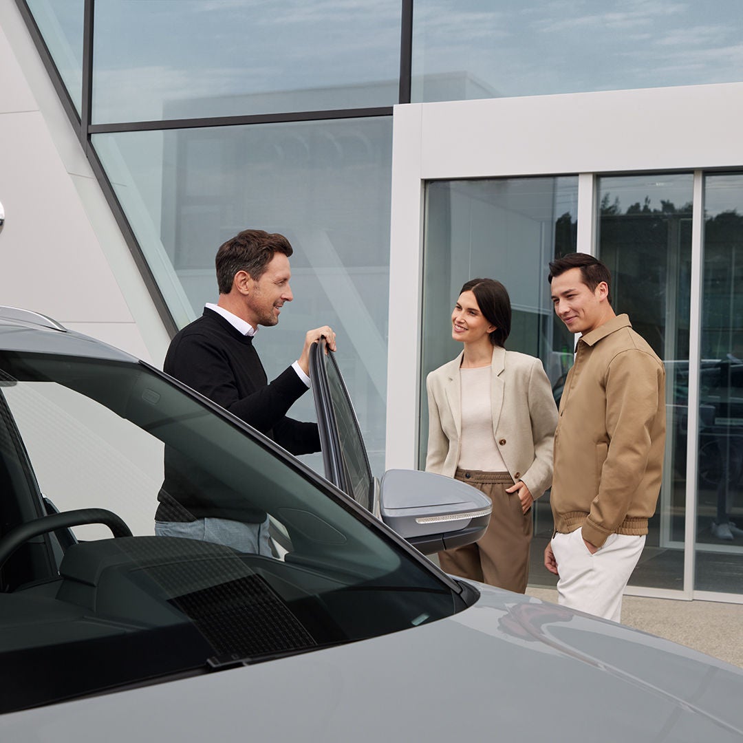 Verkäufer zeigt einem Paar einen grauen Audi vor einem modernen Gebäude mit Glasfassade, alle Personen lächeln.