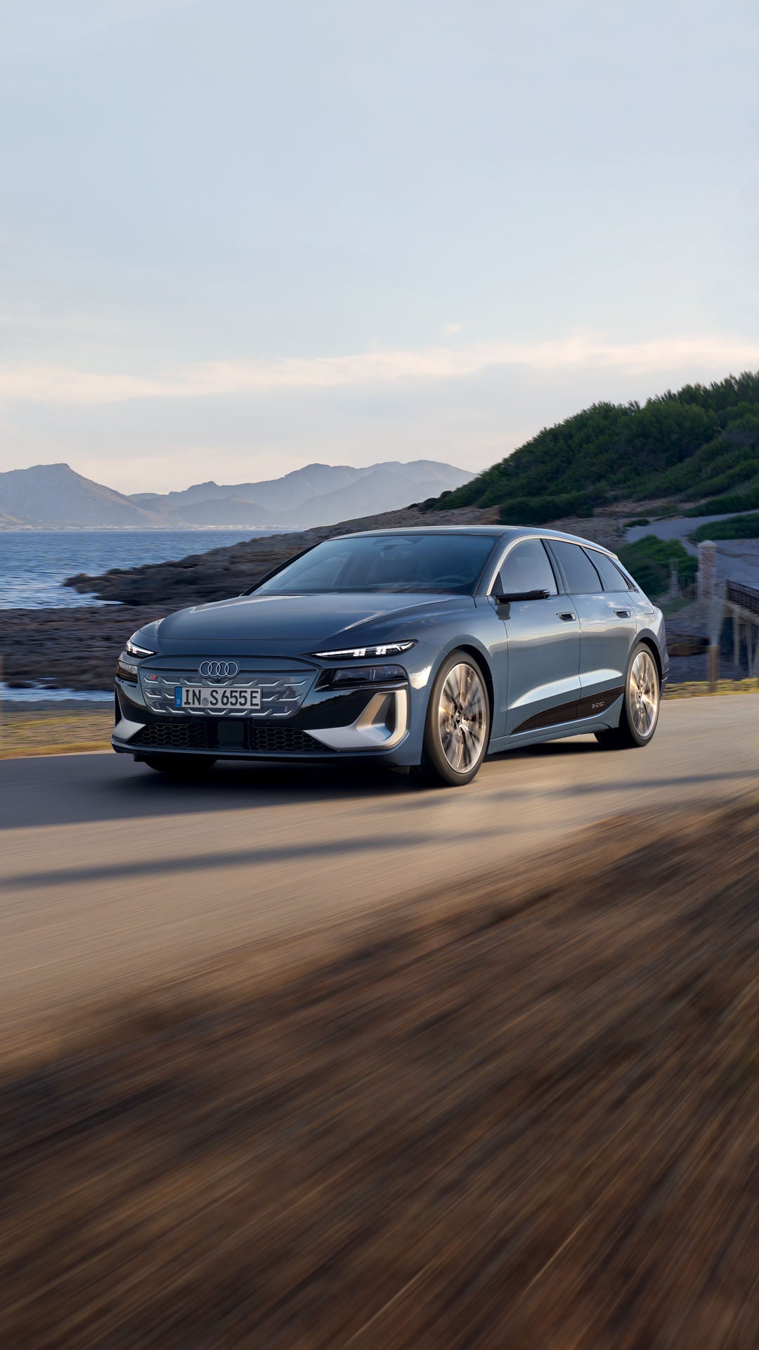 Der S6 Avant e-tron aus schräger Frontansicht, wie er eine Straße an der Küste entlangfährt. Im Hintergrund sind Berge zu sehen.