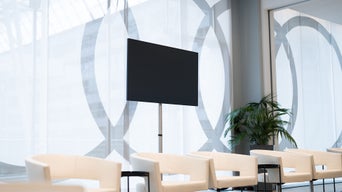 Monitor auf Stativ vor Glaswand mit Muster, helle Sessel und Pflanze im Raum.