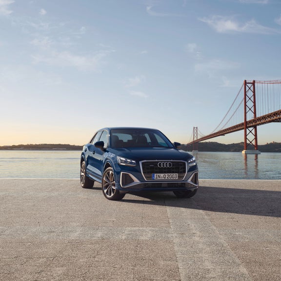 Blue car parked in front of a suspension bridge over water at sunset.