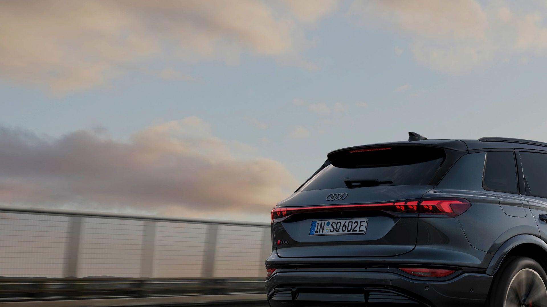 A sleek gray Audi SUV drives away on a bridge under a colorful sky, showcasing its modern design and dynamic rear lights.
