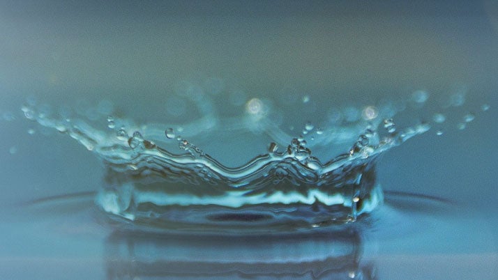 Nahaufnahme eines Wassertropfens, der auf eine Wasseroberfläche trifft und eine spritzende Krone bildet.