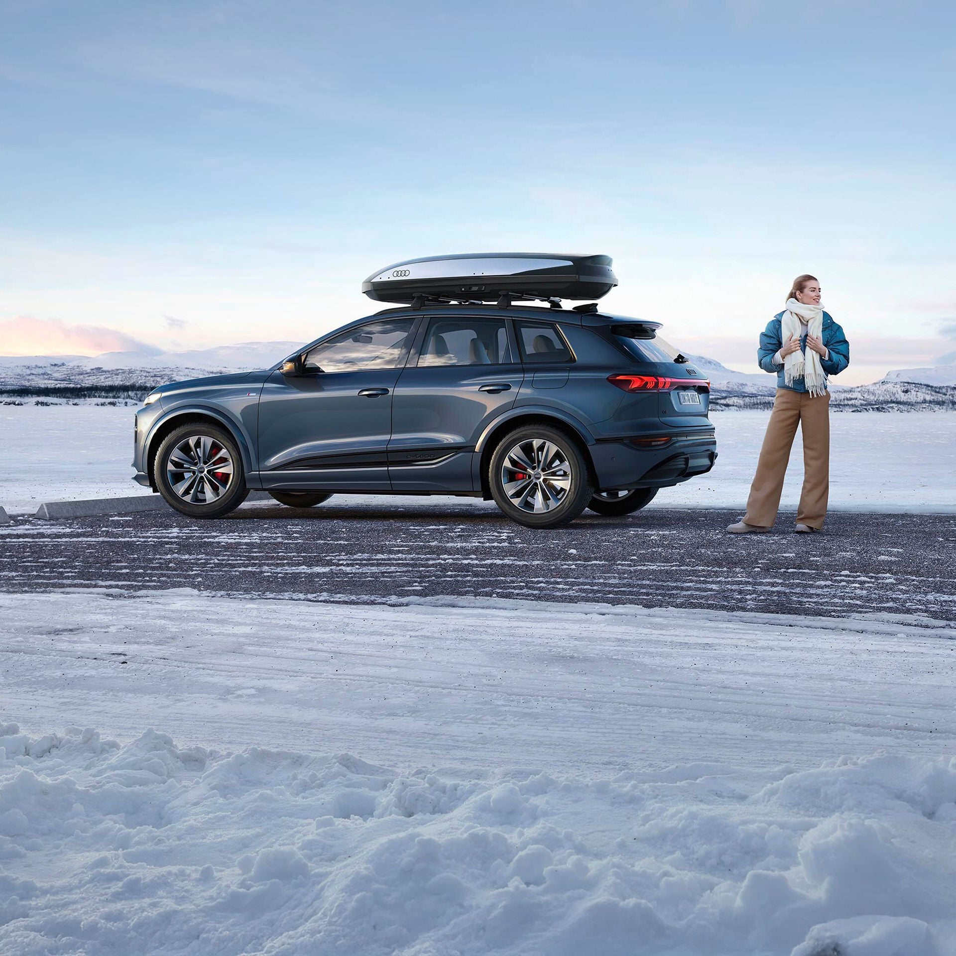 Eine Frau steht rechts neben einem blauen Audi mit Dachbox in einer Schneelandschaft