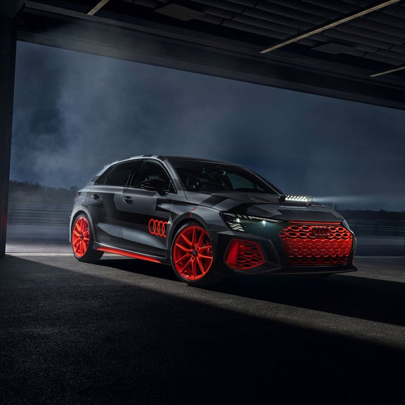 Schwarz/grauer Audi RS 3 driving experience mit orangenen Felgen und Streifendekor steht beleuchtet in dunkler Halle. 