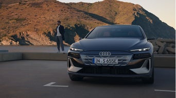 Woman in orange dress and white jacket walking beside a silver Audi RS e-tron GT car on a coastal road.