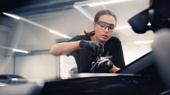 Mechanikerin mit Schutzbrille behandelt die Frontscheibe mit elektrischem Werkzeug in Werkstatt.