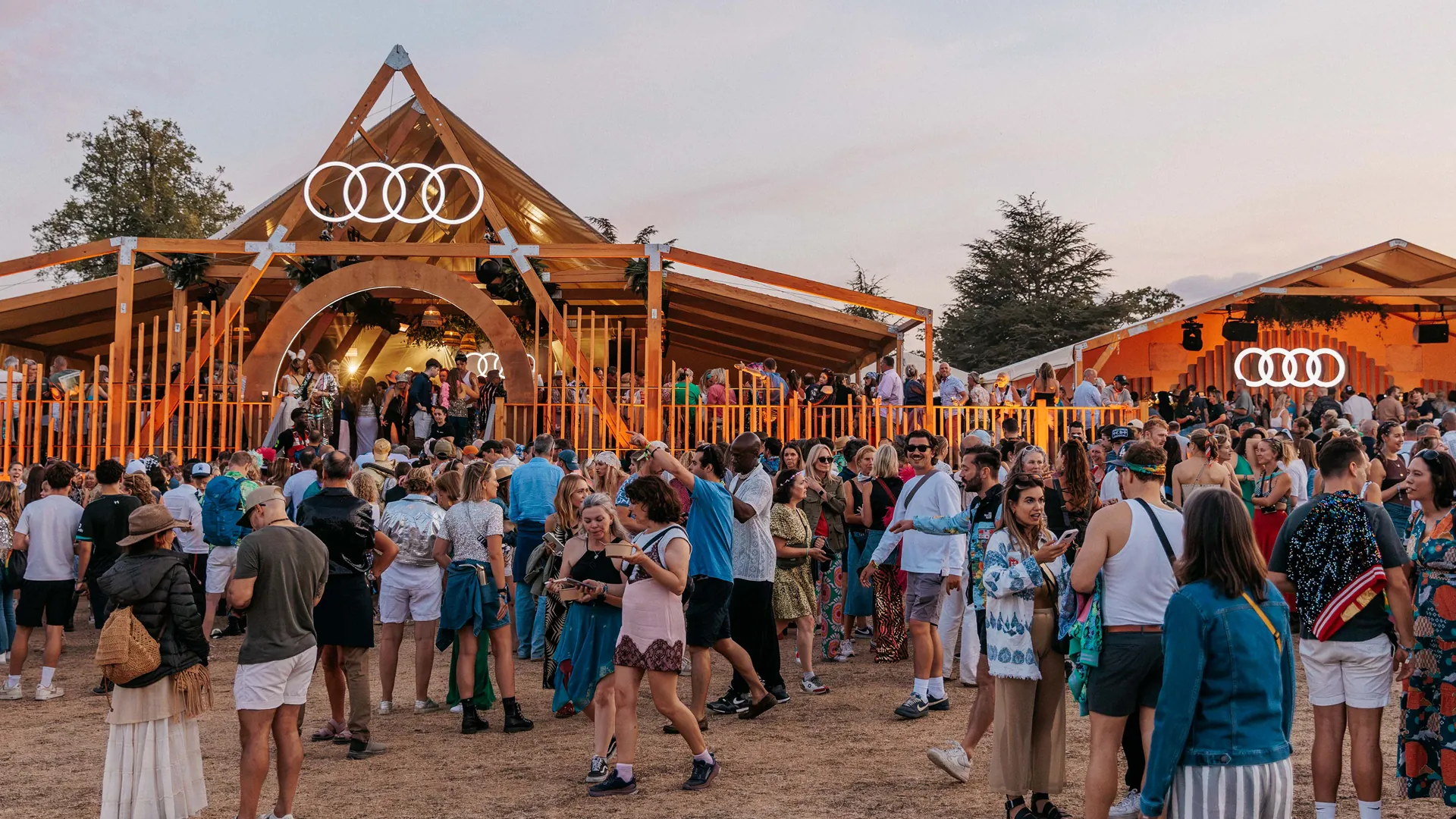Three people laugh together arm-in-arm in front of an Audi-branded festival pavilion, with a gold electric Audi and performers in shimmering gold costumes nearby. Three people laugh together arm-in-arm in front of an Audi-branded festival pavilion, with a gold electric Audi and performers in shimmering gold costumes nearby.