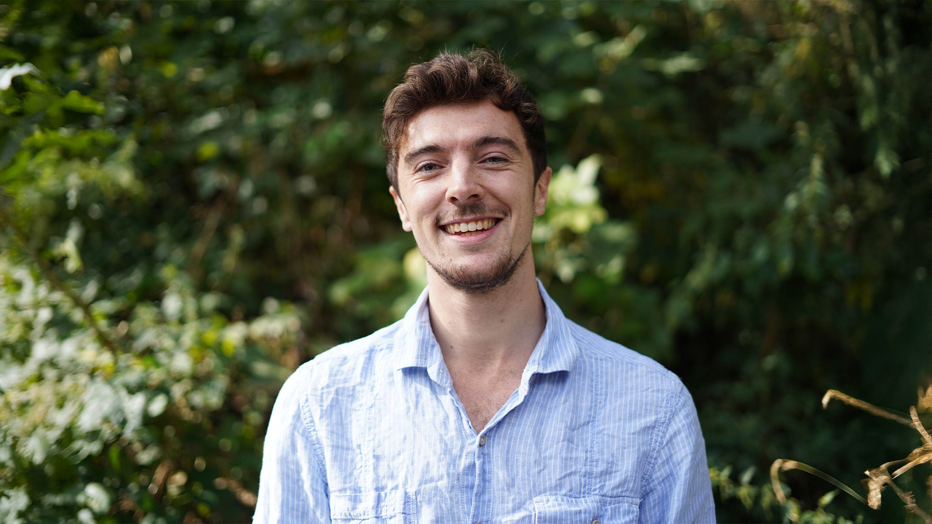 Porträt eines jungen Mannes im hellblauen Hemd, lächelnd, vor grünem, unscharfem Hintergrund.