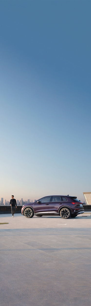 Lila Audi-SUV lädt an Ladesäule auf offenem Platz, ein Mann geht vorbei, im Hintergrund eine Skyline bei Sonnenaufgang.