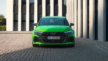 Der RS 3 Sportback von vorn parkend auf Kopfsteinpflaster, im Hintergrund Umrisse eines modernen Gebäudes.