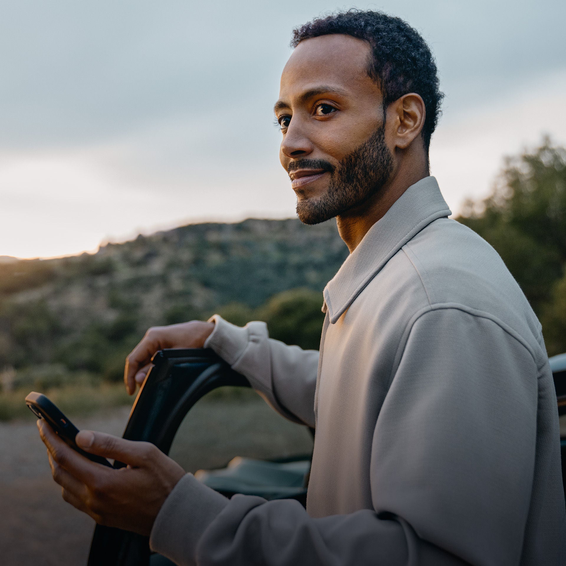 A man with a mobile phone in his hand leans gently smiling against the open driver's door of a vehicle in front of a wooded mountain backdrop at sunrise.