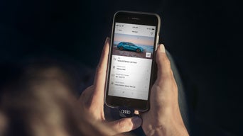 Person hält Smartphone mit geöffneter Audi-App, auf dem Display sind Fahrzeugdaten und ein blaues Audi Modell zu sehen.