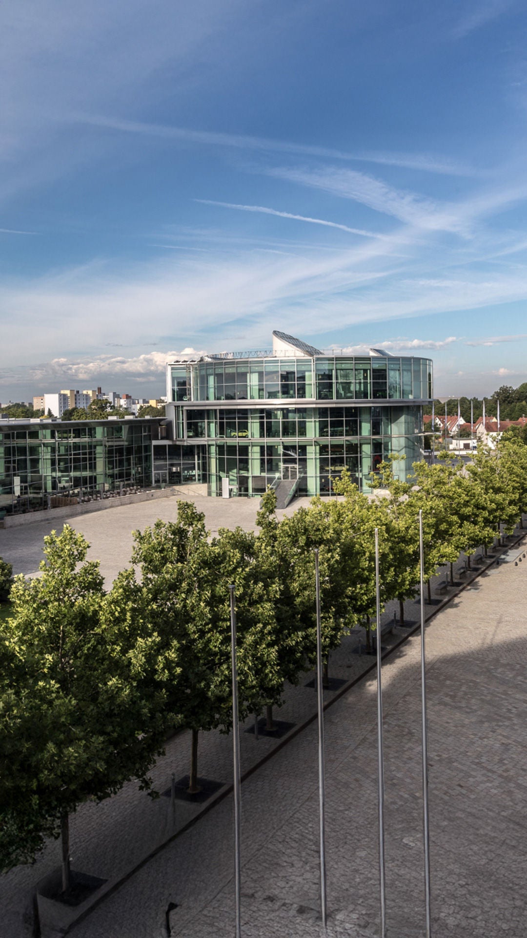 Ein moderner Gebäudekomplex mit großen Glasfassaden und geschwungenen Formen, umgeben von einer Baumreihe und gepflasterten Wegen unter einem klaren blauen Himmel. 