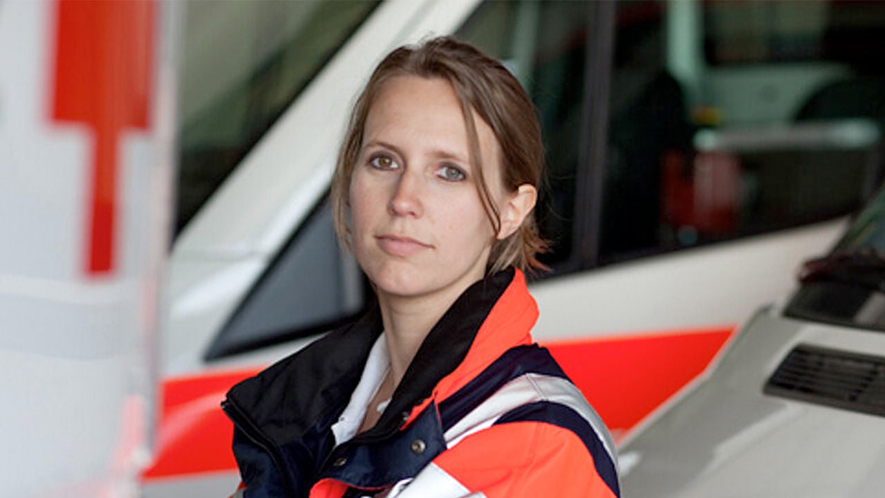 Frau in Einsatzkleidung steht vor einem Rettungswagen mit rotem Kreuz, Blick in die Kamera, Hintergrund unscharf.