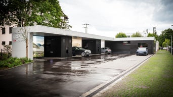 Audi Charging Hub München Westkreuz bei Tag mit weißer Fassade, Audi-Logo und Grünflächen rechts, im Hintergrund Wohngebäude.