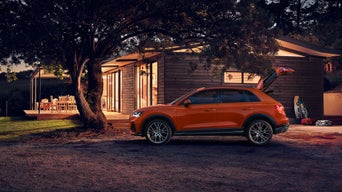 Ein orangefarbener SUV steht bei Dämmerung vor einem beleuchteten Holzhaus, umgeben von Bäumen und Freizeitgeräten.