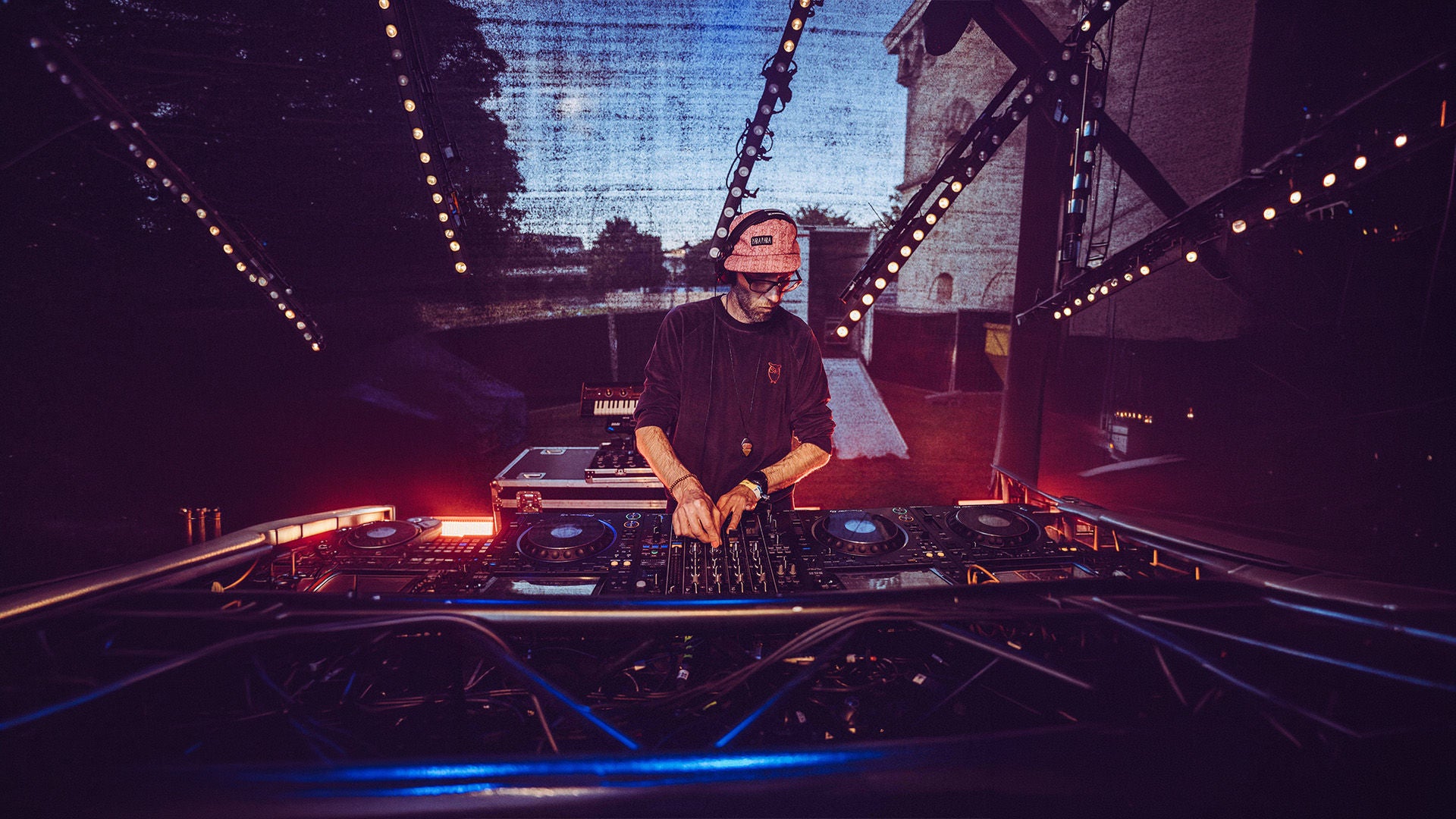 DJ mit Cap und Brille bedient ein großes Mischpult auf einer Bühne im Freien, umgeben von warmen LED-Lichtern.