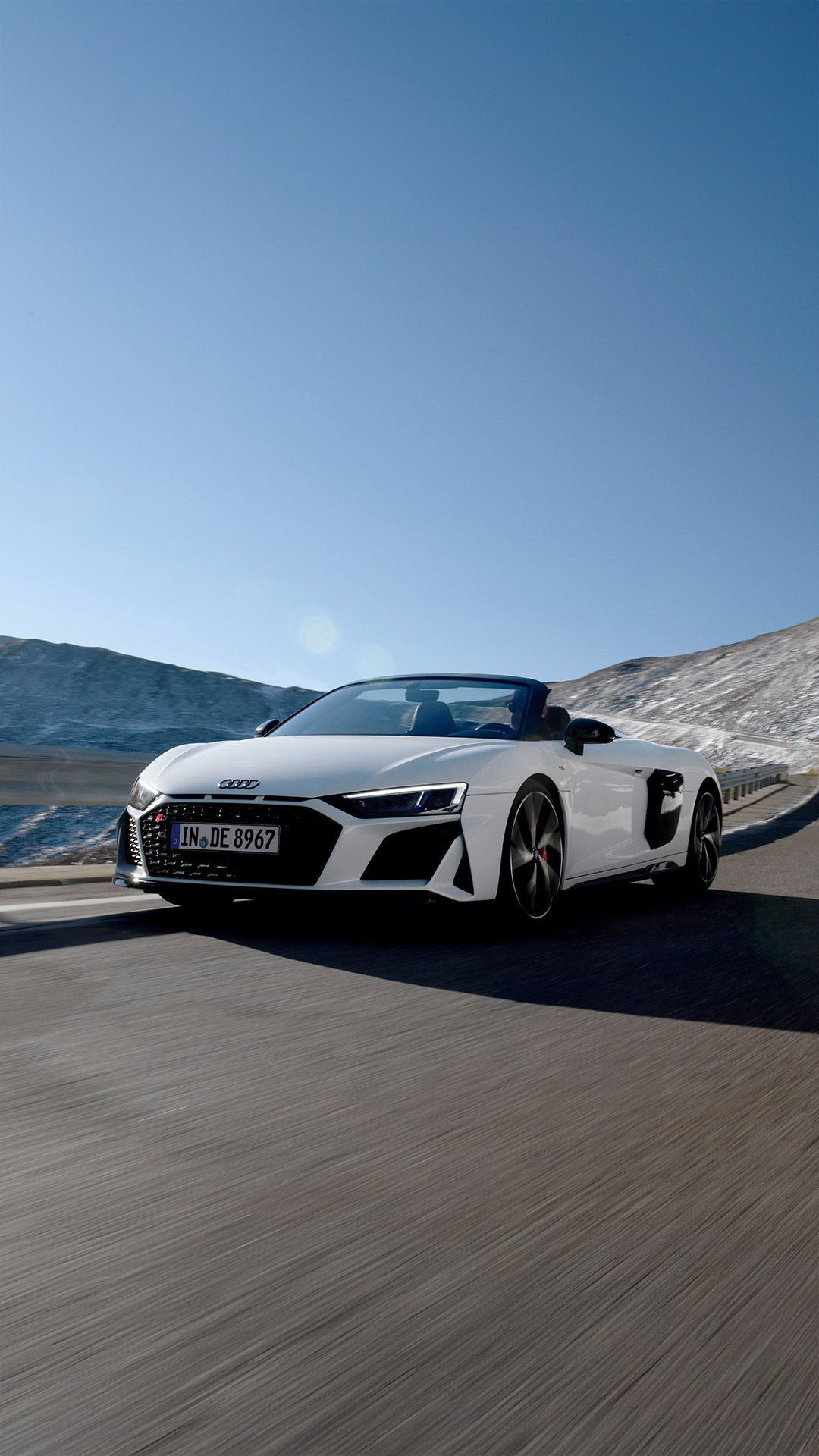 Weißer Audi R8 Spyder V10 performance quattro fährt in den Bergen bei sonnigem Wetter mit blauem Himmel.