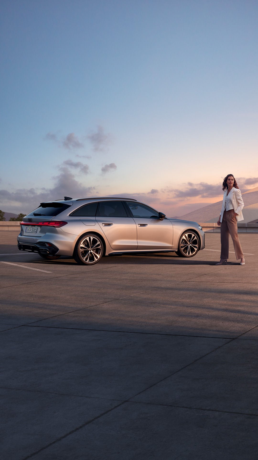 Eine Frau steht neben einem silbernen Audi Modell bei Sonnenuntergang und guckt in die Ferne