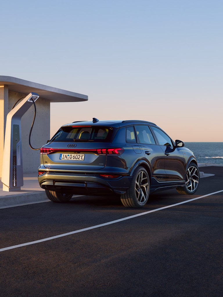 Ein Audi Q6 SUV e-tron wird an einer Ladesäule auf einem Parkplatz geladen, im Hintergrund das Meer bei Sonnenuntergang.