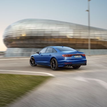 Ein blauer Audi S8 TFSI fährt auf kurviger Straße, im Hintergrund ein großes, rundes, modernes Gebäude mit Glasfassade.