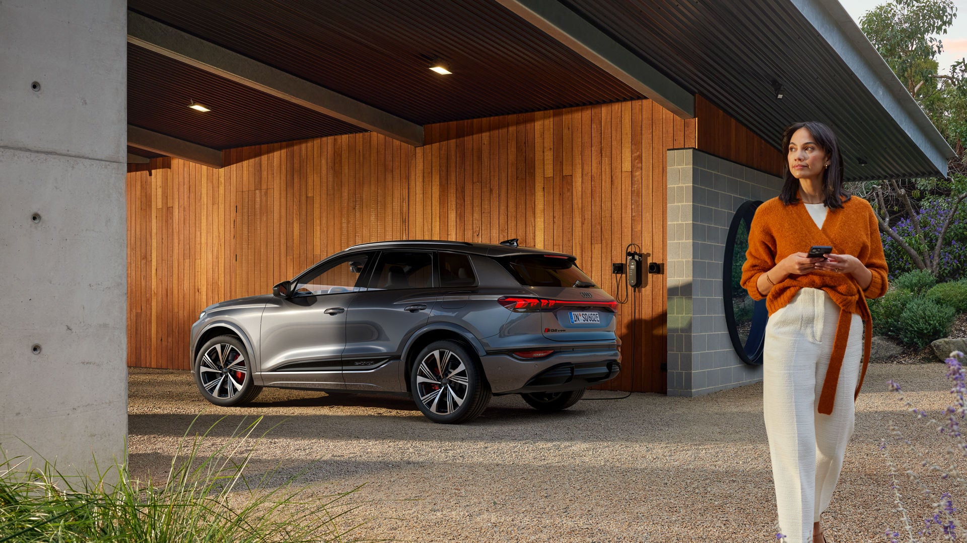 Eine Frau steht mit Smartphone neben einem silbernen Audi Q6 e-tron, der an einer Hausladestation lädt.