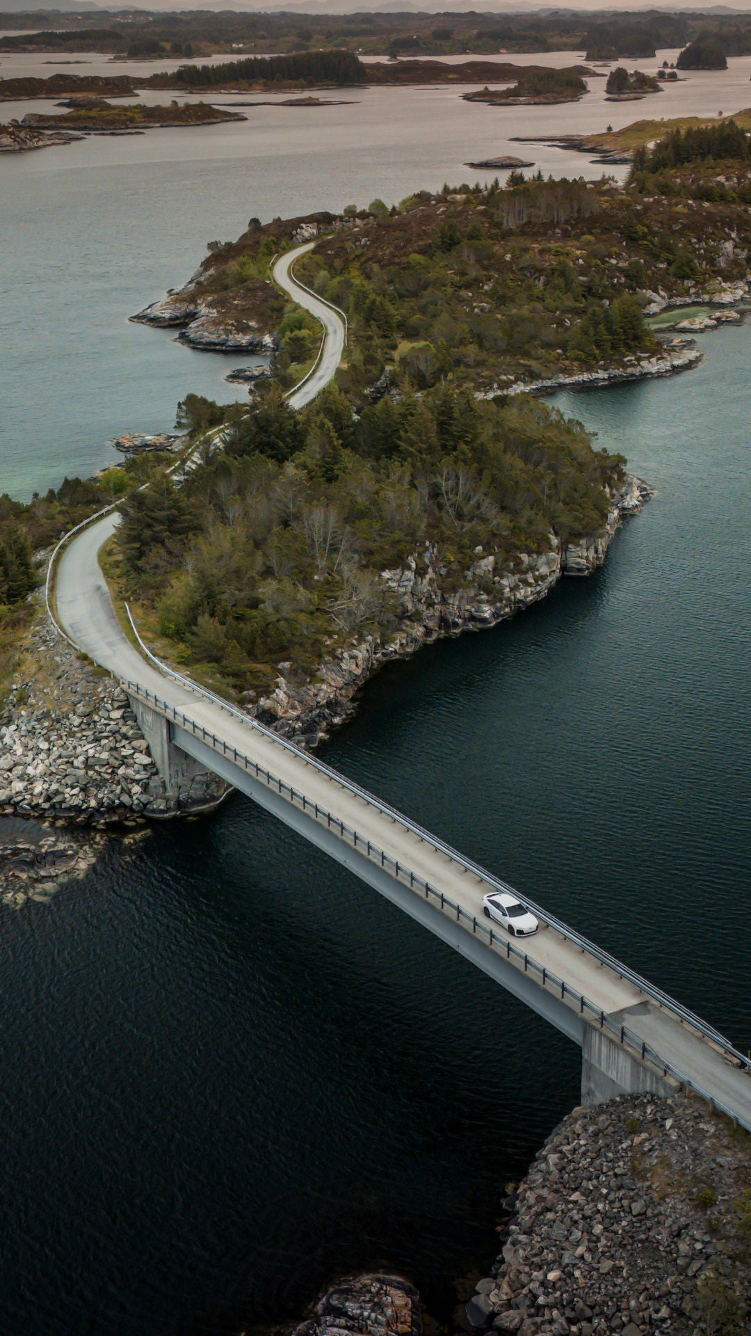 An Audi A6 Sportback e-tron in Glacier white metallic crosses a bridge in Norway’s rugged coastal landscape. There are numerous small islands in the background.