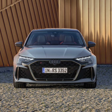 Frontansicht einer Audi RS 3 Limousine mit beleuchteten LED-Scheinwerfern vor einer modernen, kupferfarbenen Wand.