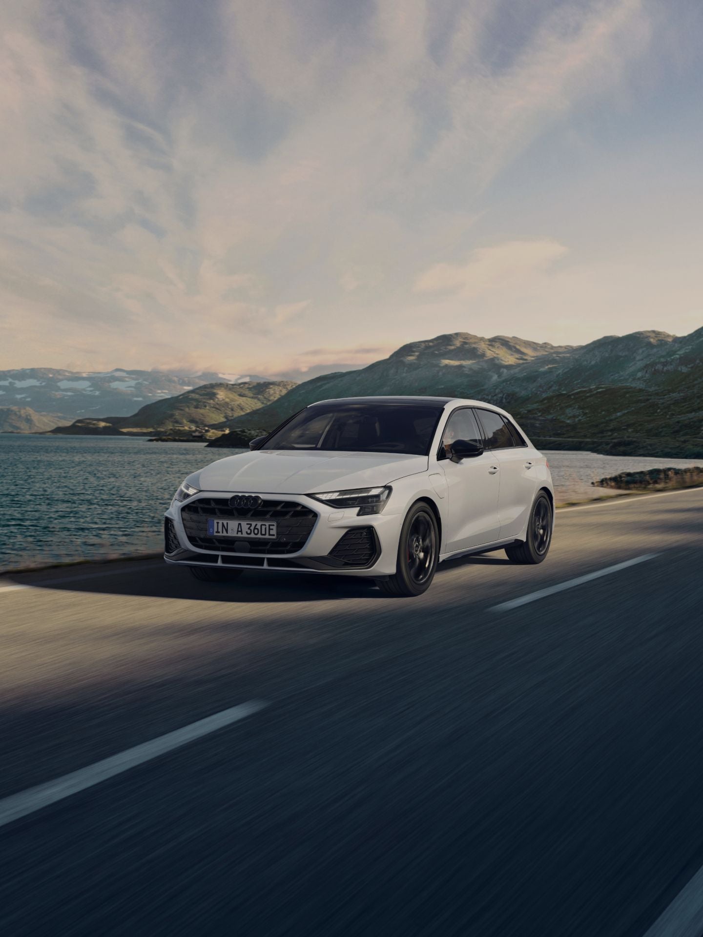White Audi car driving along a coastal road with mountains in the background.