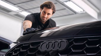 Mann poliert die Motorhaube eines blauen Audi mit Poliermaschine in heller Werkstattumgebung.