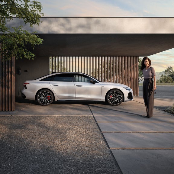 Die A6 Limousine e-hybrid steht seitlich vor einer Hauswand, an welche eine Ladestation montiert ist. Vor dem Auto steht eine junge Frau.