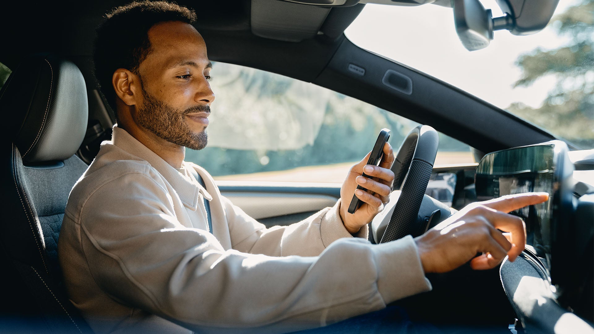 Ein Mann mit beiger Jacke sitzt auf dem Fahrersitz und tippt mit der rechten Hand auf das zentrale digitale Display im Fahrzeug und hält in der linken Hand ein Smartphone.