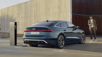 The S6 Sportback e-tron in a slanted rear profile. The vehicle is parked in front of a large building with wood panelling and is connected to a charging station. A young man stands in front of the vehicle, looking at the smartphone in his hand.