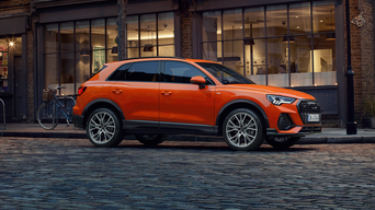 Oranger Audi Q3 steht bei Dämmerung auf Kopfsteinpflaster vor einem Café mit großen Fenstern und Fahrrad im Hintergrund, Seitenansicht.